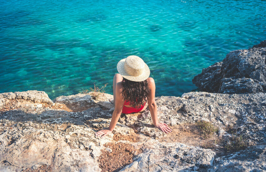 Eine Frau sitz auf einem Fels mit Blick auf das türkise Meer.